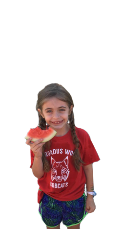 Anna, a small girl wearing a red shirt and eating a watermelon, stands and smiles.