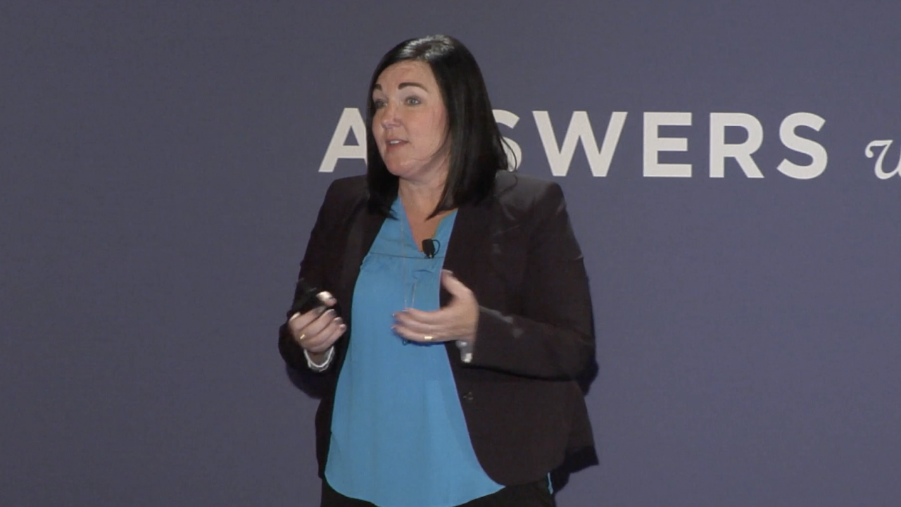 Jeanne Alhusen stands on a stage and speaks with a blue backdrop.