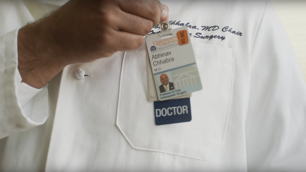 Dr. Chhabra's white coat, you see his hand hooking his medical badge to his pocket.