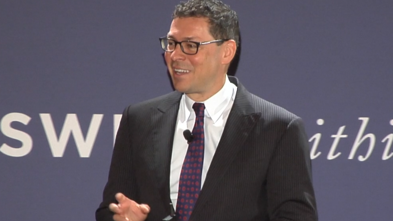 Dr. Oberholzer stands in front of a blue backdrop presenting. 