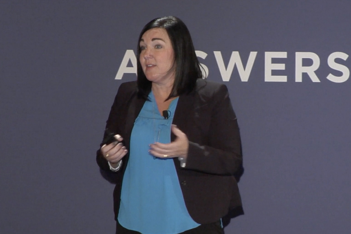 Jeanne Alhusen stands on a stage and speaks with a blue backdrop.