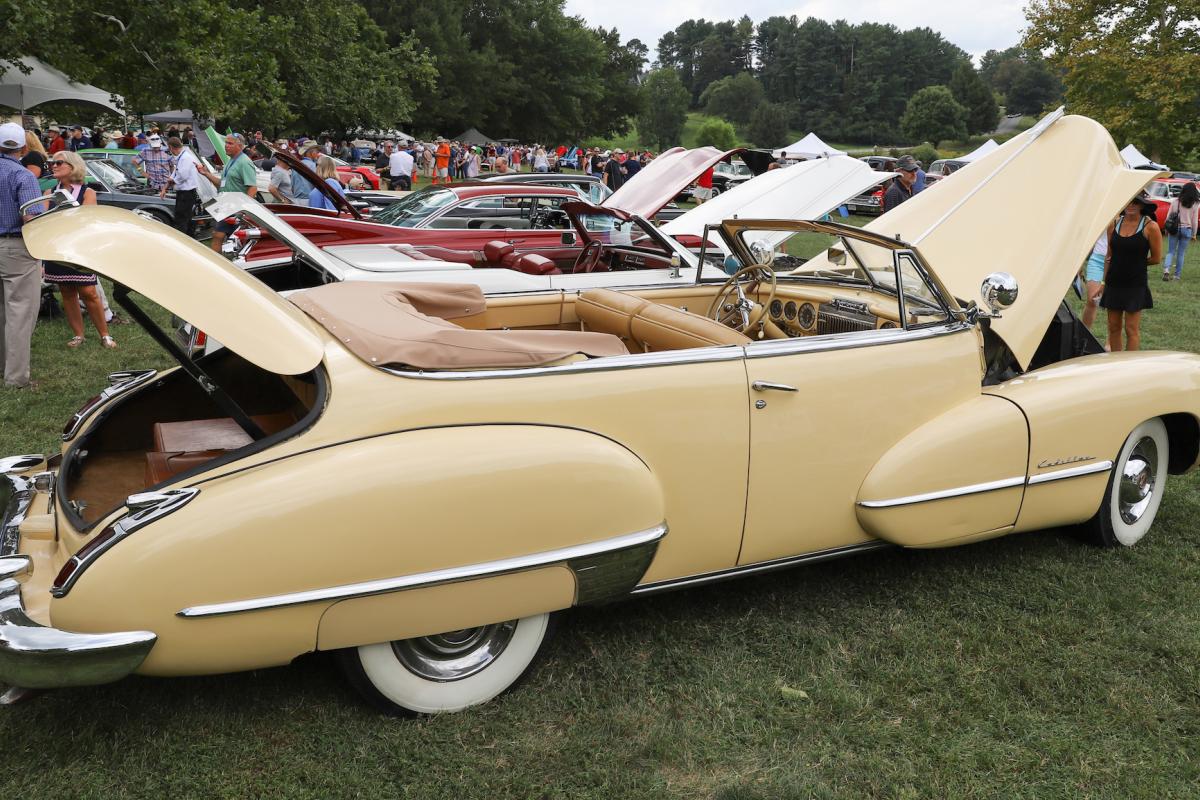 A vintage yellow car is parking with its trunk and engine popped open, crowds can be seen in the background.