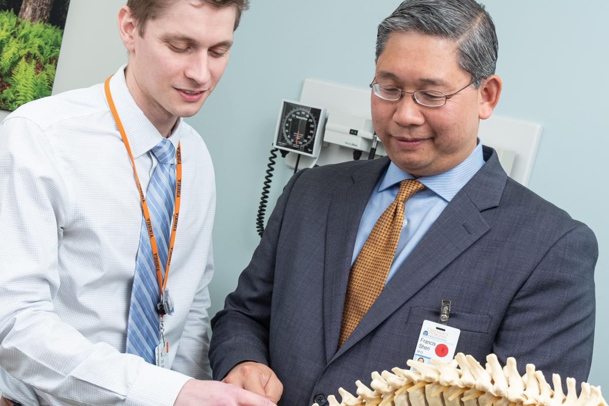 A resident stands beside Dr. Shen and they both examine vertebrae on a human skeleton spine.