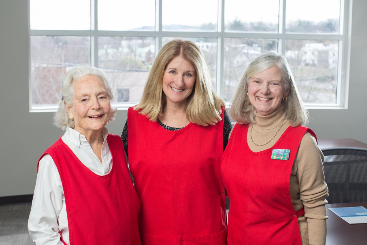 (From left to right) In their red volunteer aprons, UVA Cancer Center Advisory Board Members Ginny Semmes, Wendy Seay, and Martha Weiss, smile for a photo.